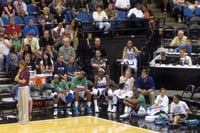 Corner of a basketball court mid-game, with fans seated in rows of tiered seats, and players sitting in chairs and on the floor below them. 